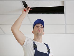 man in white t-shirt and blue overalls looking under a ceiling panel